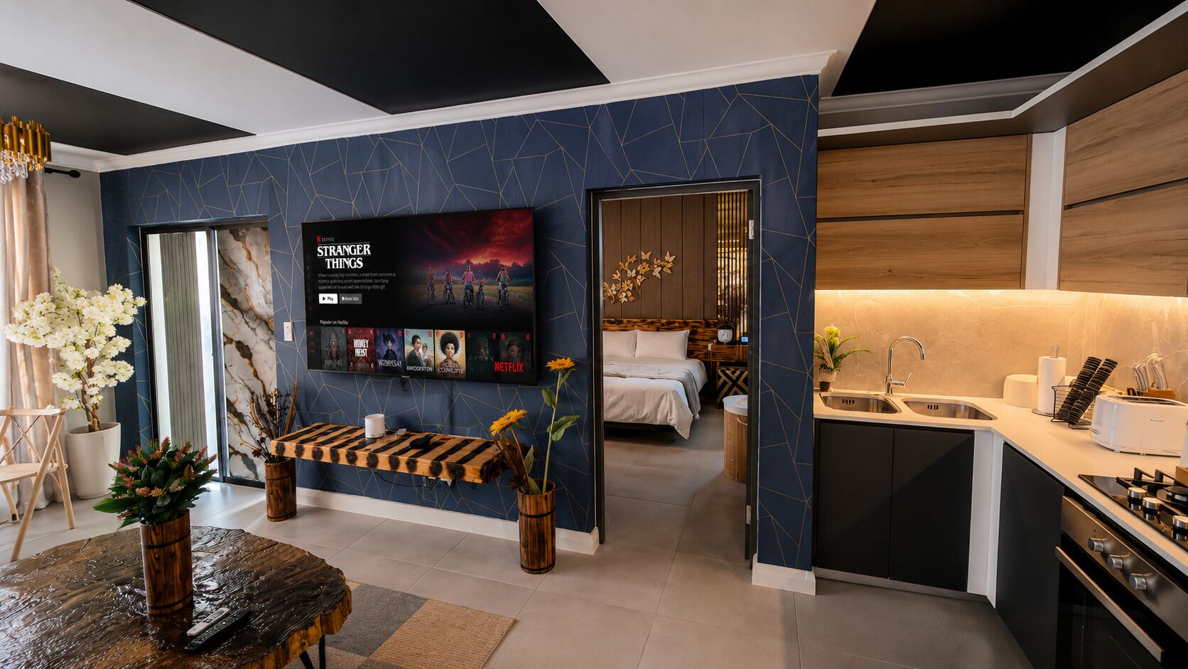 A modern interior with a TV displaying a movie, a bed, a kitchen area, and a seating area with a coffee table and plants.