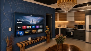 Ein modernes Wohnzimmer mit Flachbildfernseher, der einen Fernsehsender zeigt, einer Holzbank und einer Küchenzeile mit Edelstahlgeräten.