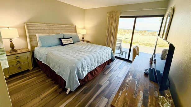 A hotel room with a bed, bedside lamps, a wooden floor, a sliding glass door to the beach, and a television. - Steps from the Beach with Gulf View Ground Floor (Fort Walton Beach)