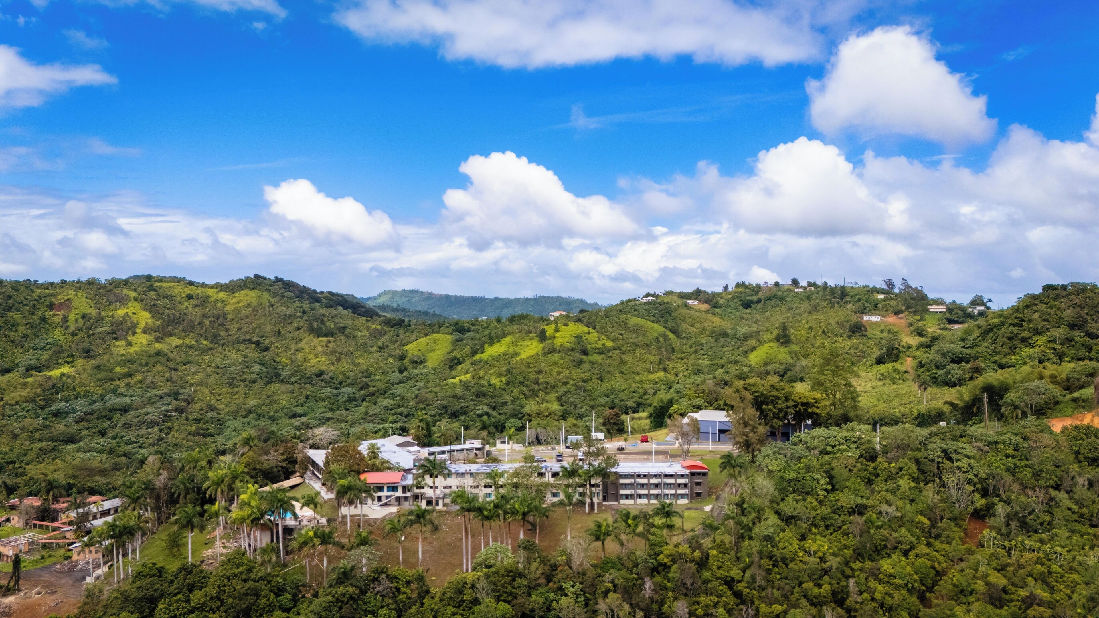 Un paisaje montañoso con un pequeño conjunto de edificios que incluye varias construcciones, rodeado de vegetación exuberante y un cielo despejado.