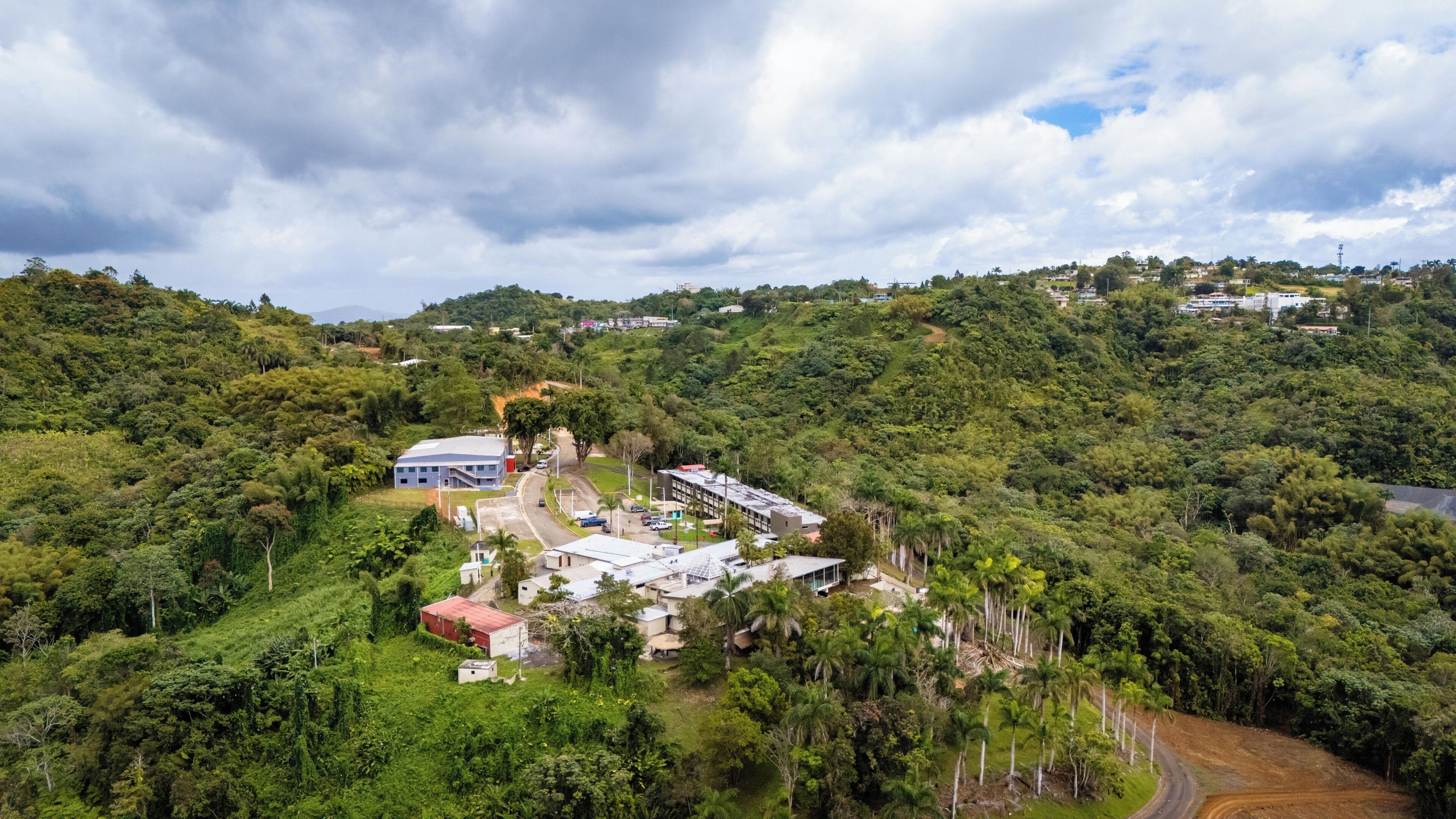 Una zona montañosa con un pequeño grupo de edificios, incluyendo una gran estructura azul y varios edificios más pequeños, rodeada de abundante vegetación.