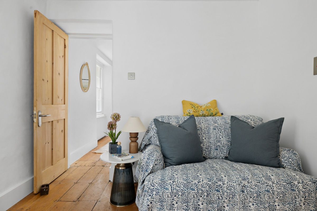 A room with a wooden door, a small round table with a lamp, a sofa with patterned upholstery, and a yellow pillow.