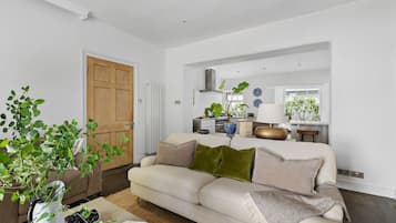 Ein modernes Wohnzimmer mit einem beigen Sofa, einem Couchtisch mit Obst und Brot, einer Zimmerpflanze und Blick auf eine Küche und einen Essbereich.