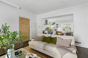 A modern living room with a beige sofa, a coffee table with fruits and bread, a potted plant, and a view of a kitchen and dining area. - Lantern House (Torquay)