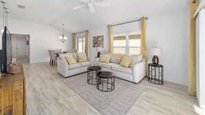 A spacious living room with a white sofa set, two side tables, a ceiling fan, and a dining area in the background. - Walk to Brownwood! Spacious 3BR | Golfcart + BBQ (The Villages)