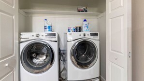 A laundry room with two front-loading washing machines and shelves above containing detergent bottles. - Lux Modern Getaway At&t Stadium Views King Bed (Arlington)