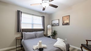 A hotel room with a bed, a ceiling fan, a window with curtains, a bedside table, a chair, and two wall-mounted pictures. - Lux Modern Getaway At&t Stadium Views King Bed (Arlington)