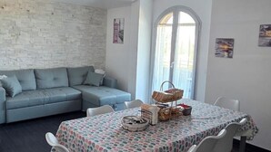 A room with a sectional sofa, a dining table with a patterned tablecloth, a window with curtains, and wall art. - Apartment in Villa Gra&Mary (Tombaccia)