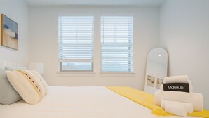 Un dormitorio con cama, una silla, un espejo y dos ventanas con persianas.
