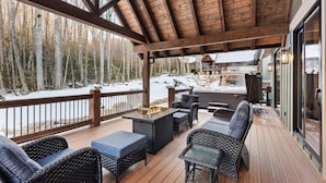 A covered outdoor deck with wicker furniture, a fire pit, and a view of a snowy forest. - The Cabins on Buckeye Creek at Eagles Nest (Banner Elk)