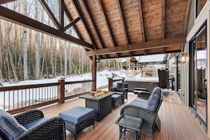 A covered outdoor deck with wicker furniture, a fire pit, and a view of a snowy forest. - The Cabins on Buckeye Creek at Eagles Nest (Banner Elk)