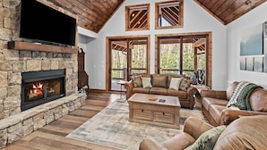 A cozy living room with a stone fireplace, leather sofas, a flat-screen TV, and large windows with wooden frames. - The Cabins on Buckeye Creek at Eagles Nest (Banner Elk)
