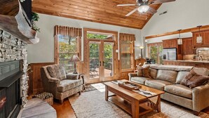 Ein gemütliches Wohnzimmer mit Kamin, Deckenventilator, Sofa, Couchtisch und einer Küchenzeile mit Holzausbau und Kühlschrank.