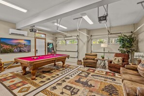 Ein Billardtisch, eine Couch, eine Lampe und ein Fernseher in einem Raum mit Fenstern und Deckenventilator.