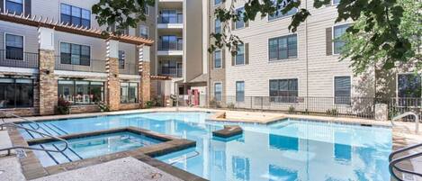 A swimming pool surrounded by multi-story buildings with balconies, a covered patio area, and a clear sky.