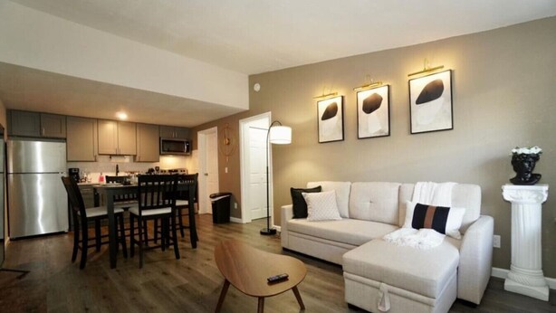 A modern living room with a sofa, coffee table, and dining area with chairs and a kitchen in the background. - King Suite - 10 min to Ft Moore (Columbus)