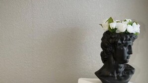 A room with a beige sofa, a white cushion, and a black vase with white flowers on a white pedestal. - King Suite - 10 min to Ft Moore (Columbus)