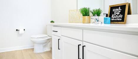 Un baño moderno con inodoro, armarios blancos, una alfombra a rayas y un letrero motivacional sobre la encimera.