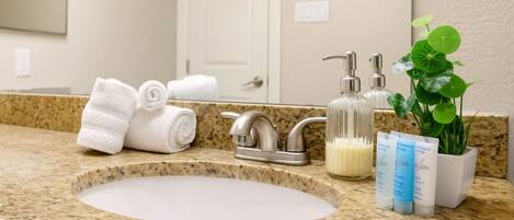 A bathroom with a granite countertop, a sink, a mirror, a towel rack, a soap dispenser, and a potted plant.