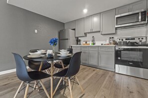 Una cocina moderna con armarios grises, un refrigerador de acero inoxidable, un microondas y un horno. Hay una mesa de comedor con sillas negras y un florero con flores azules.