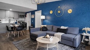 Una sala moderna con una pared azul de acento, un sofá gris, una mesa de centro redonda y una zona de comedor con una mesa y sillas blancas.