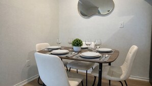 A dining area with a table set for four, white chairs, a potted plant, and a wall-mounted mirror. - 2713 Kathrynln2 Plano 1BR (Plano)