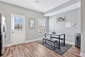 A room with a door, two windows with blinds, a table with a bench, a mirror, and a trash can.