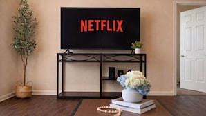 Un salon avec un téléviseur affichant le logo « NETFLIX », une plante, un vase rempli de fleurs et une petite table sur laquelle se trouvent un livre et un collier de perles.