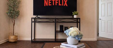 Sala de estar con televisor mostrando 'NETFLIX', una planta, un jarrón con flores y una mesita con un libro y un collar de cuentas.