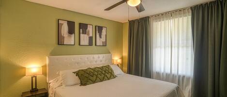 A bedroom with a tufted headboard, a bed with a white quilt, a green throw pillow, a ceiling fan, and a window with curtains.