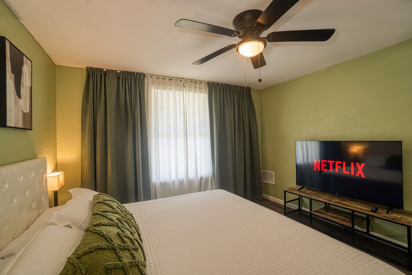 A hotel room with a bed, a ceiling fan, a TV, and a window with curtains.