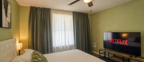 A hotel room with a bed, a ceiling fan, a TV, and a window with curtains.