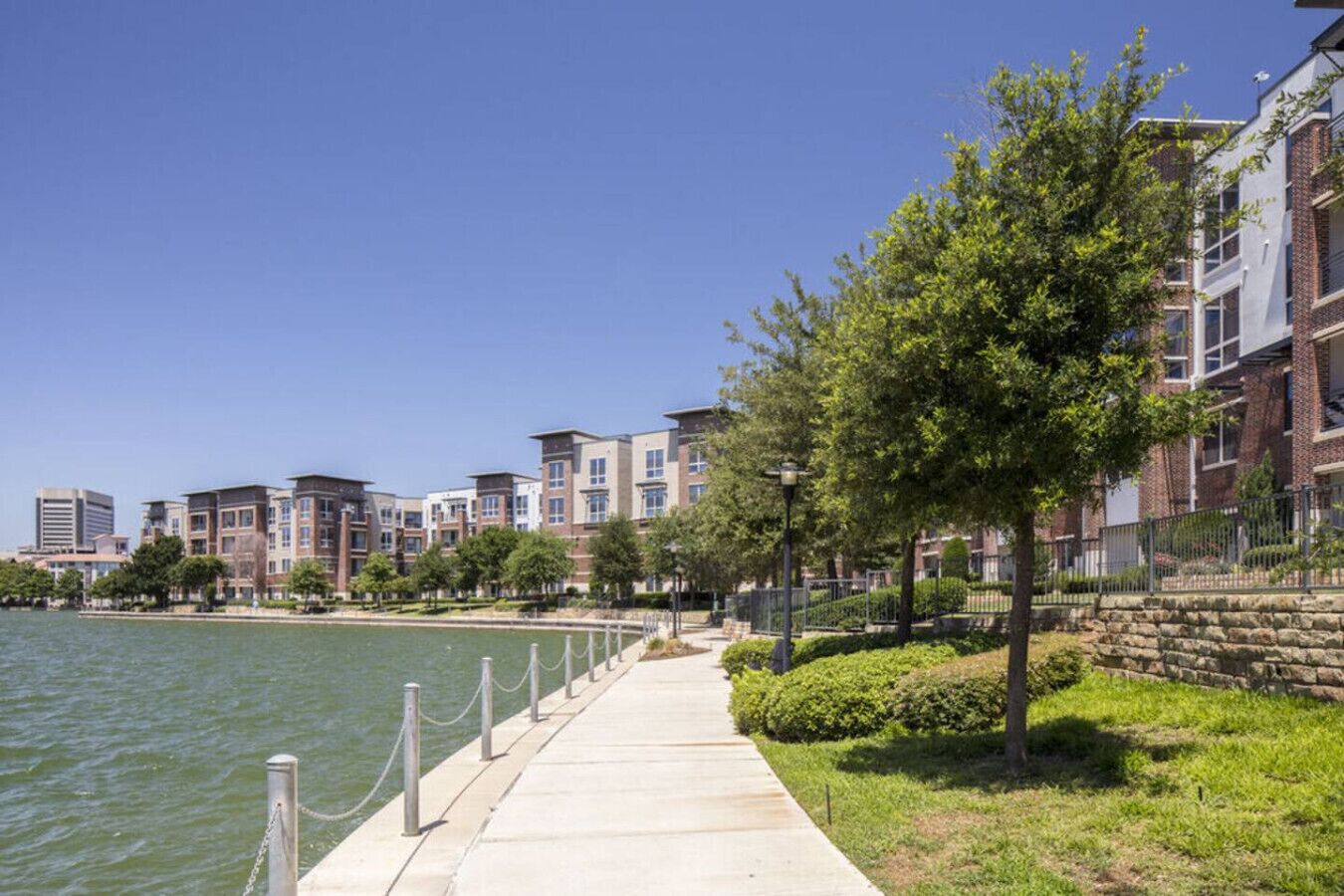 A waterfront promenade with a concrete walkway, trees, and residential buildings.