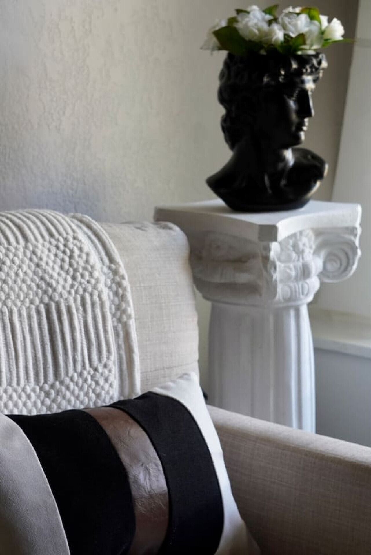 A white armchair with a textured throw blanket and a black cushion. A white vase with flowers is on a white pedestal with a classical design.