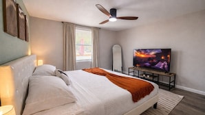 Un dormitorio con cama, ventilador de techo, televisión y ventana con cortinas.