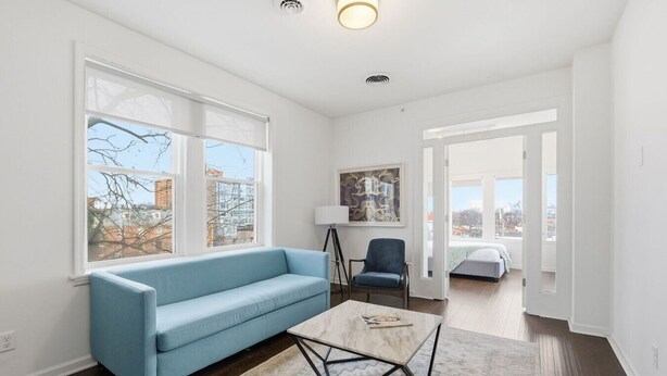 Uma sala de estar moderna com um sofá azul, uma mesa de centro e uma cadeira. Há grandes janelas com vista para a cidade, um quadro na parede e uma porta que dá para outro cômodo.