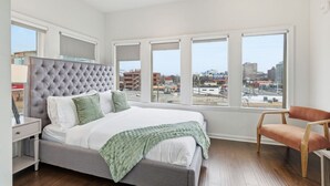Quarto com uma cama grande, uma cadeira e várias janelas com vista para a cidade.