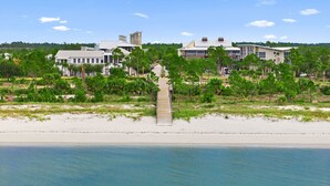 Una propiedad frente a la playa con un edificio blanco destacado, un camino de madera que lleva a la arena y abundante vegetación.