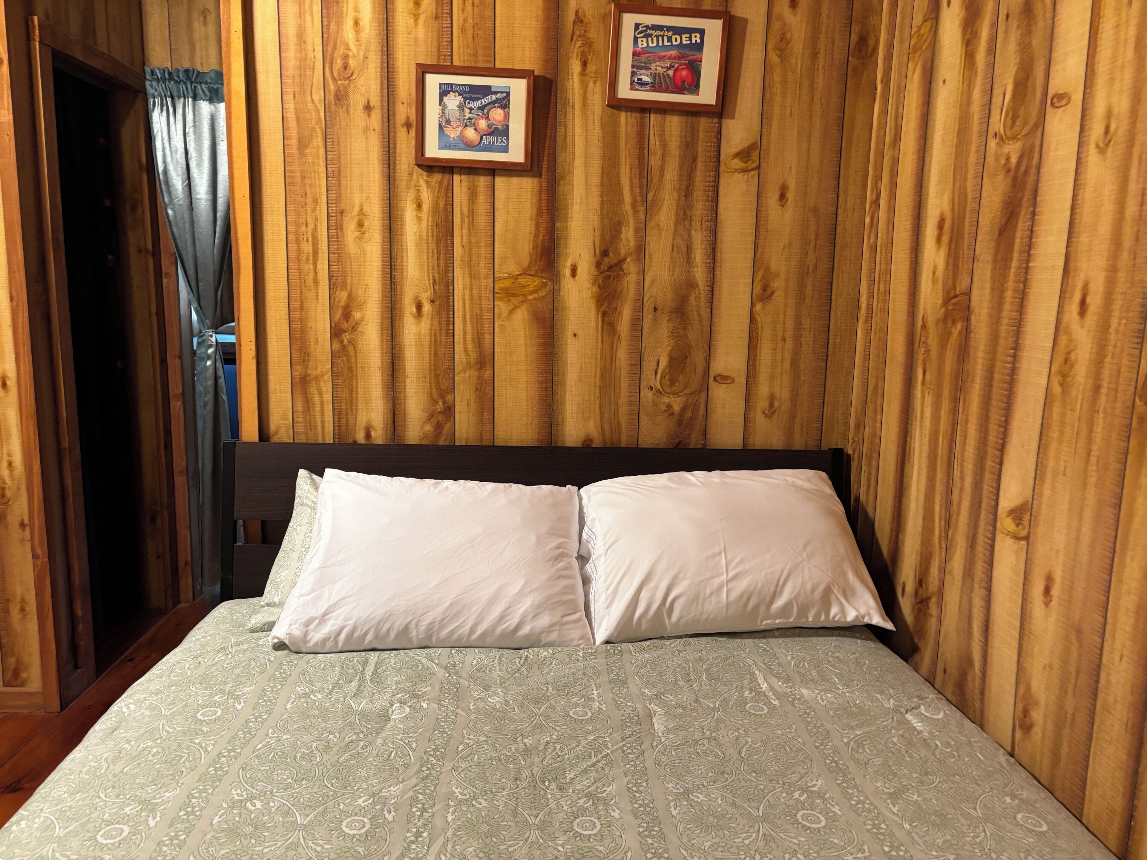 Una habitación con paneles de madera, una cama, dos cuadros en la pared y una cortina.