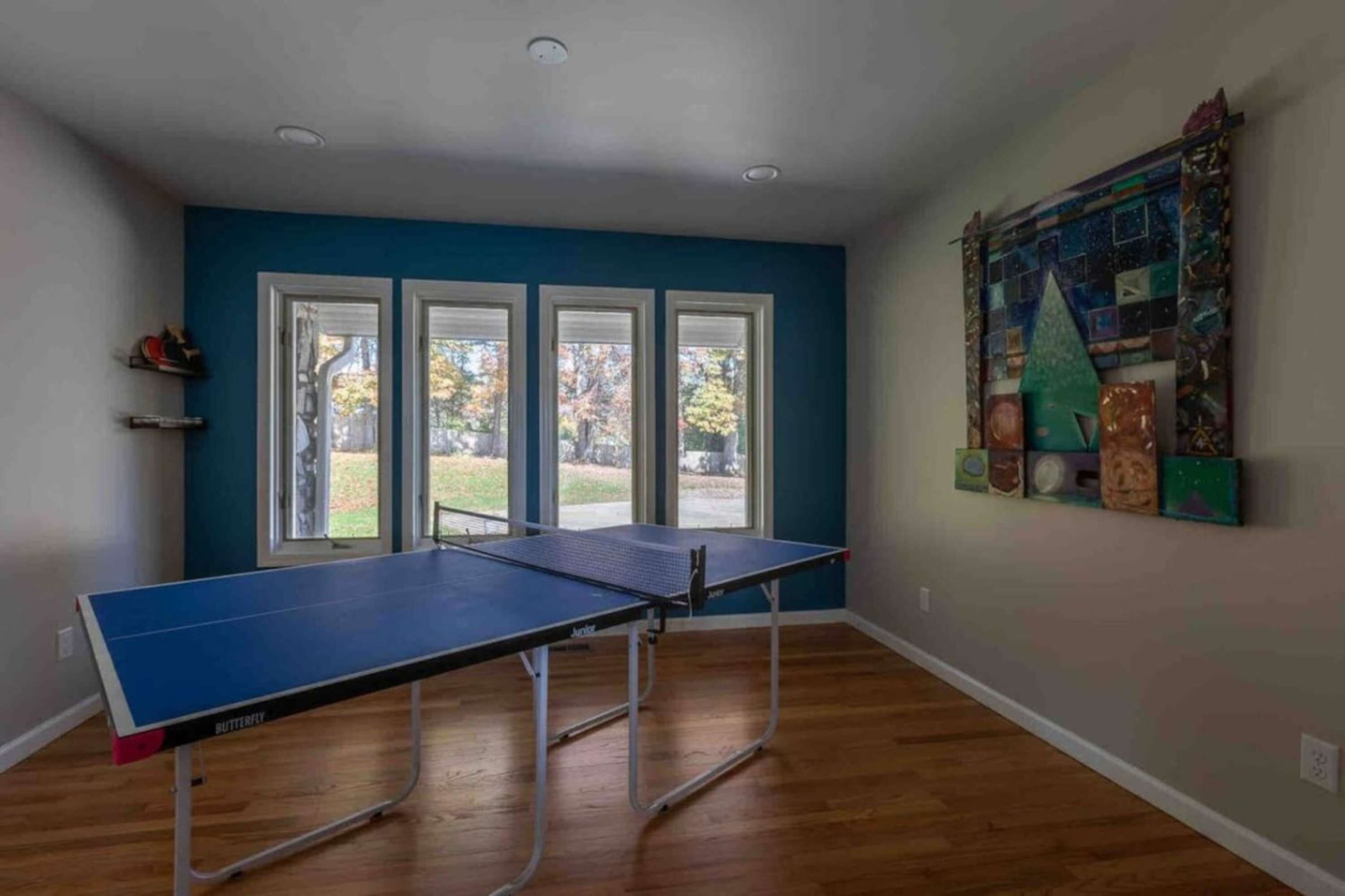 A room with a ping pong table, large windows, and a wall art piece.