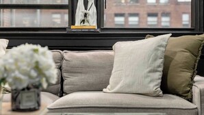 A sofa with multiple cushions, a vase with flowers, and books on a table in front of a window with a view of buildings. - Centrally Located APT in NYC Walk to Time Square (New York)