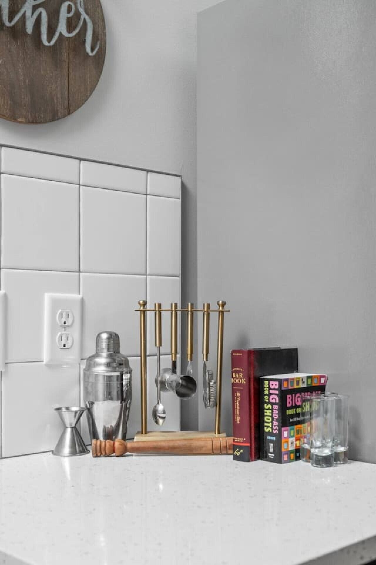 A kitchen counter with a white countertop, a wooden cutting board, a cocktail shaker, a set of gold-colored utensils, and two books titled 'Big Bad Ass' and 'Book of Shots'.
