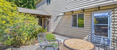 A patio with a round table and two chairs, a small garden with various plants, and a house with a window and a door.