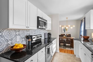 A modern kitchen with white cabinets, a black countertop, and a patterned backsplash. There is a dining area with a wooden table and chairs, and a chandelier.