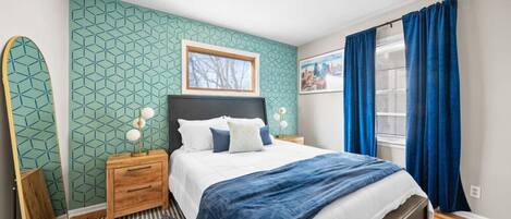 Quarto moderno com parede estampada em padrão geométrico, cama com manta azul, mesinhas de cabeceira de madeira e cortina azul.