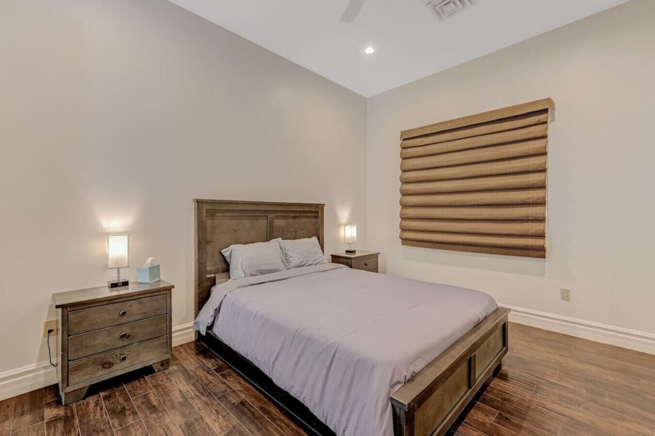 Une chambre à coucher avec un lit en bois, deux tables de chevet avec des lampes, une grande fenêtre avec des stores et un plancher en bois.