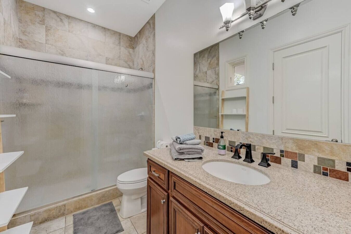 A modern bathroom with a glass-enclosed shower, a white toilet, a vanity with a sink and mirror, and a tiled backsplash.