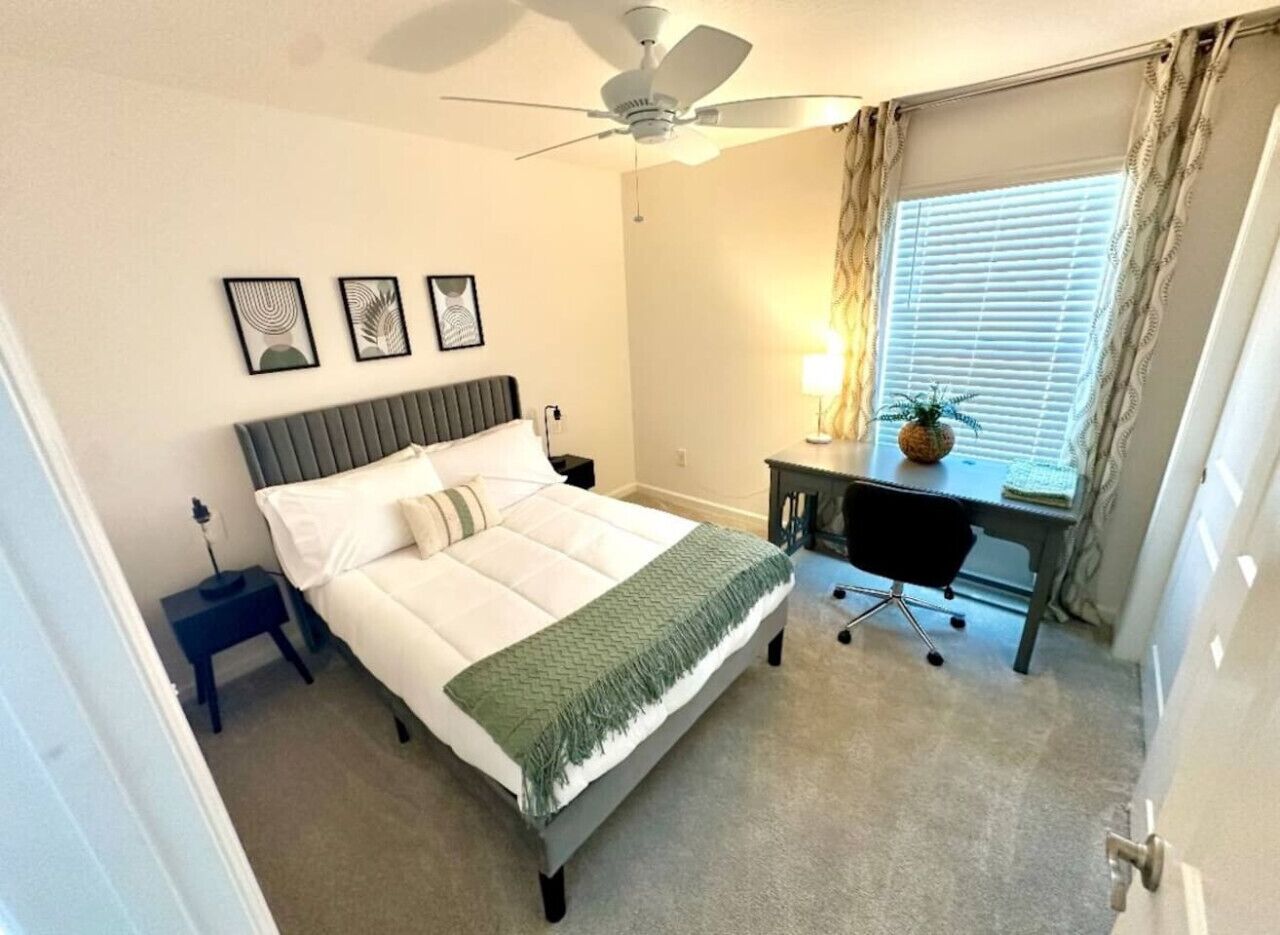 Un dormitorio con cama, escritorio, silla, ventilador de techo y ventana con cortinas.