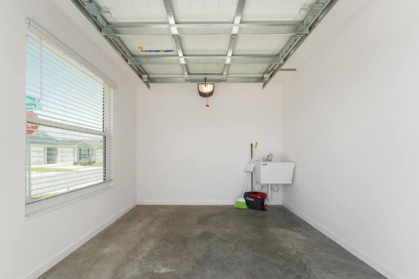 A room with a concrete floor, a window with blinds, a ceiling fan, and a mop and bucket against the wall.