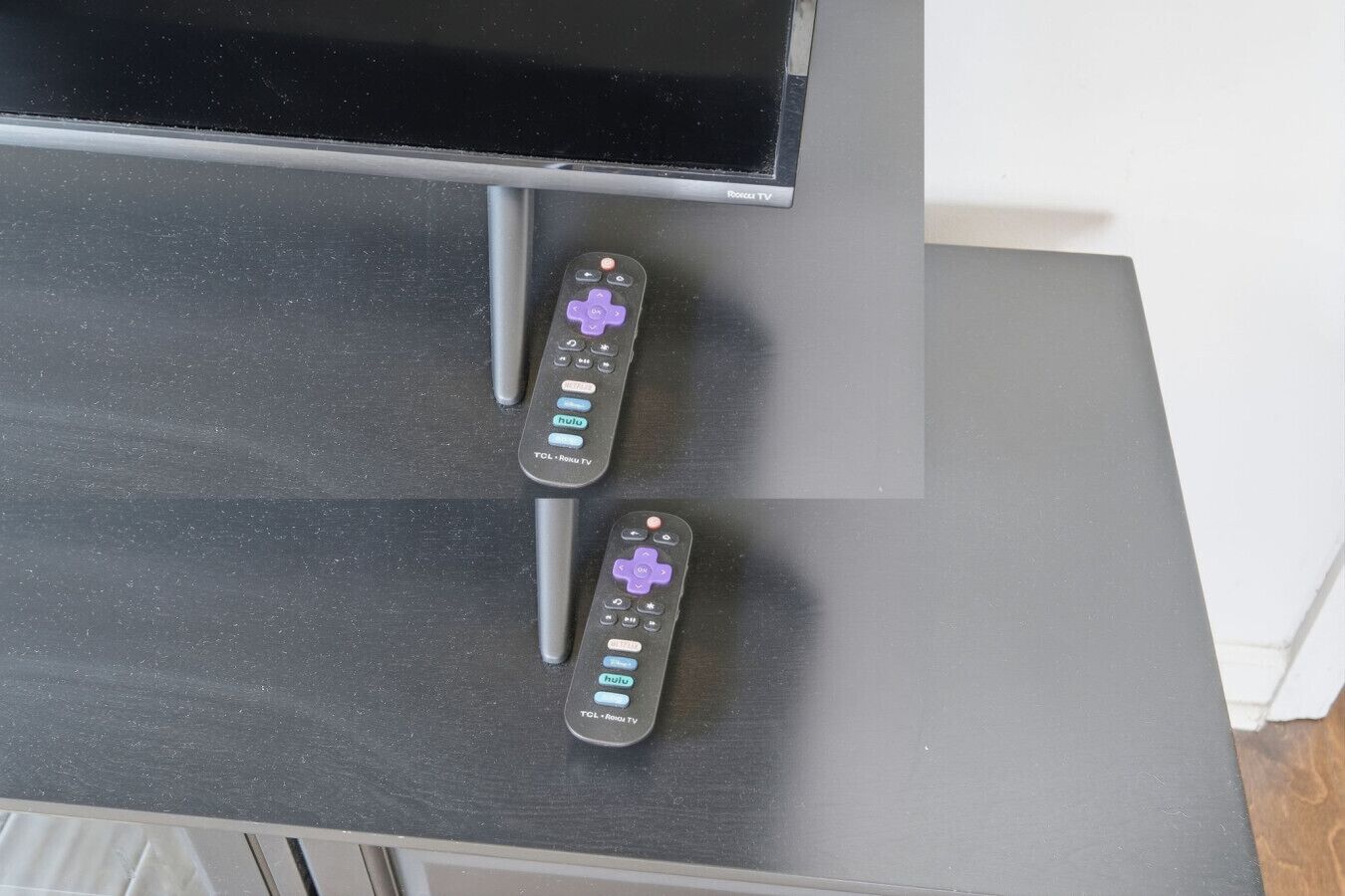 Dos controles remotos sobre una superficie negra, probablemente un mueble para televisión, con un televisor arriba.